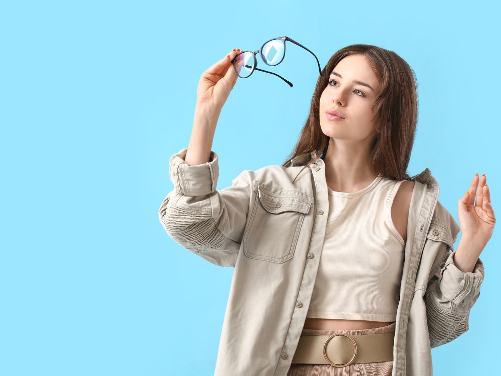 Mulher jovem analisando seus óculos contra fundo azul, ilustrando a dúvida se óculos de descanso precisa de exame.