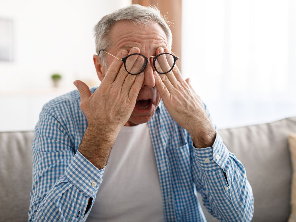 Homem mais velho com óculos esfregando os olhos cansados, mostrando sintomas que levantam a questão se óculos de descanso precisa de exame.