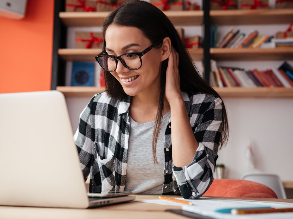 Mulher sorridente usando óculos enquanto trabalha no computador, contexto comum para avaliar se óculos de descanso precisa de exame.