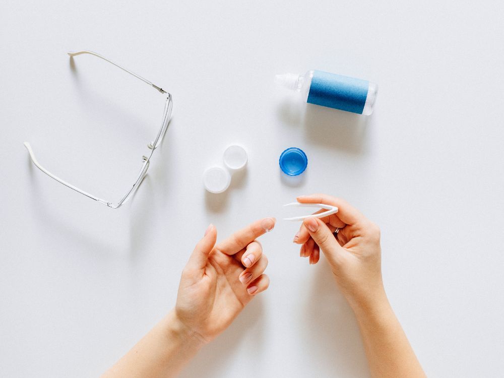 Pessoa segurando lente de contato com pinça ao lado de óculos e solução de limpeza, representando cuidados e Sintomas na adaptação de lentes de contato.